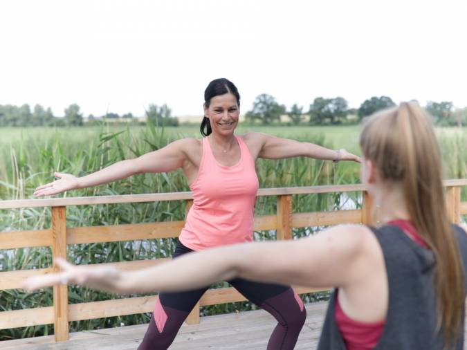 Zwei Frauen praktizieren Sport auf einer Holzterrasse, umgeben von Natur, als Teil ihrer Sporttherapie.